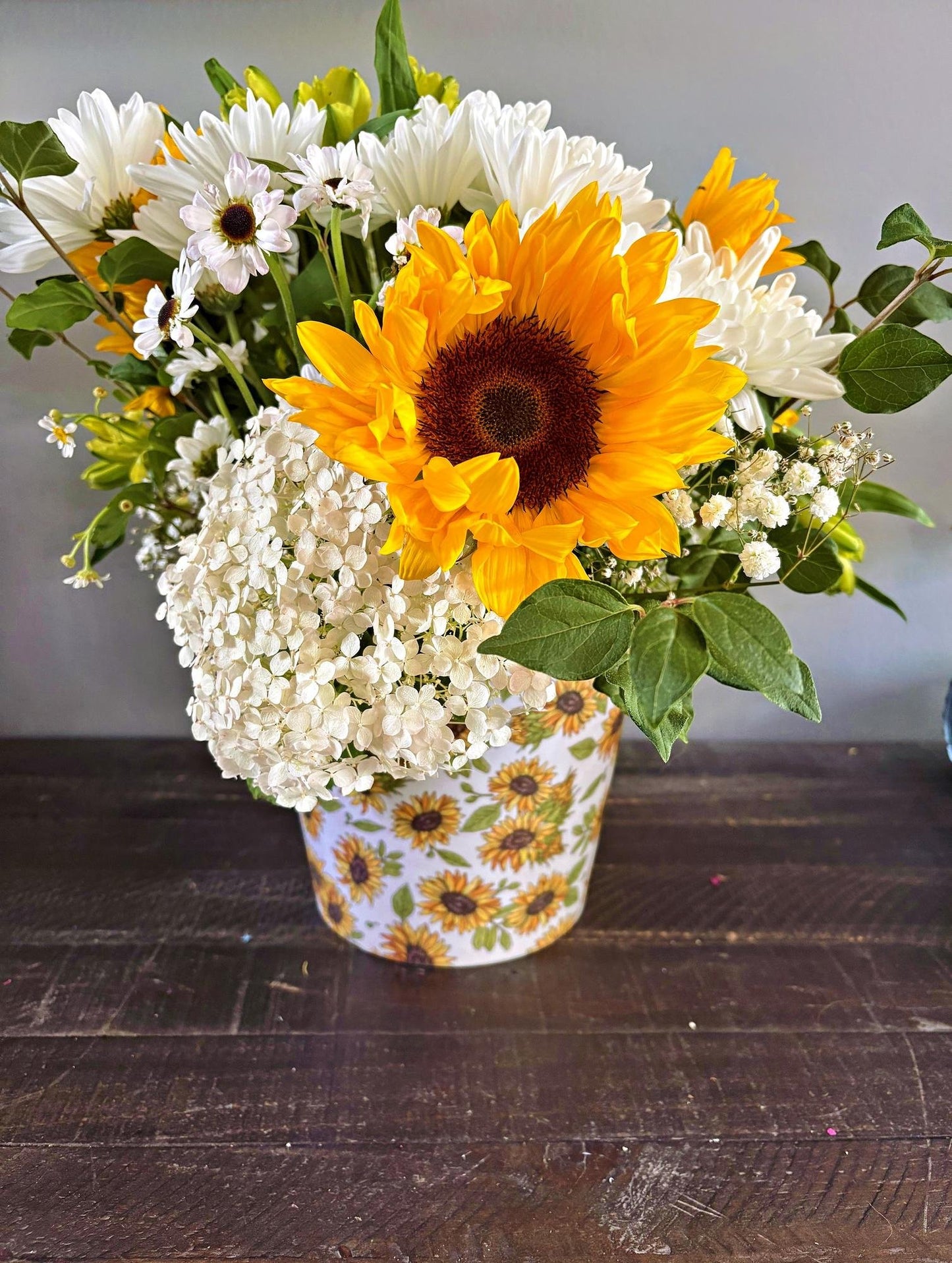 A floral arrangement featuring a large yellow sunflower, white daisies, white baby's breath, and green foliage in a white bucket decorated with a yellow sunflower pattern, set on a dark wooden table.