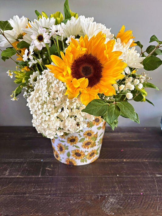 A floral arrangement featuring a large yellow sunflower, white daisies, white baby's breath, and green foliage in a white bucket decorated with a yellow sunflower pattern, set on a dark wooden table.