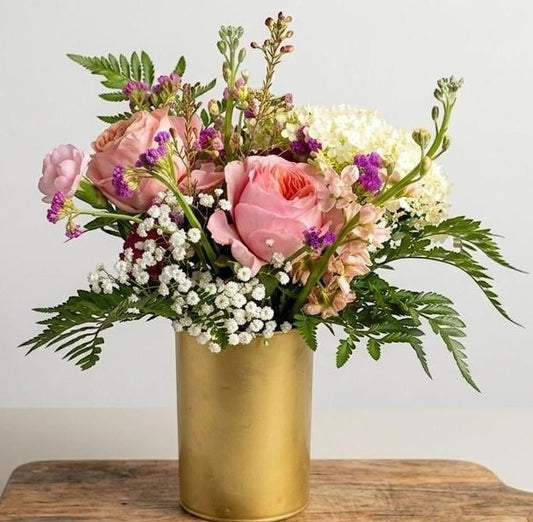 A floral arrangement featuring pink roses, white baby's breath, purple accents, and green foliage in a cylindrical gold vase, set on a wooden surface against a plain light background.